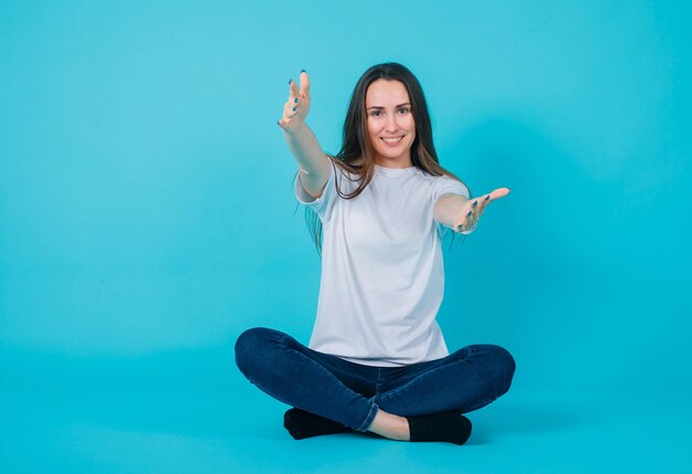 Niña sonriente está extendiendo las manos a la cámara sentándose en el suelo sobre fondo azul.