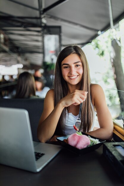 Niña sonriente está sentada en la cafetería y trabaja en su tarea en la computadora portátil durante el día