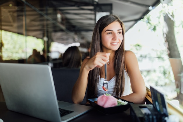 Niña sonriente está sentada en la cafetería y trabaja en su tarea en la computadora portátil durante el día