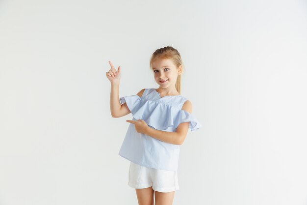 Niña sonriente con estilo posando en ropa casual aislado en la pared blanca. Modelo de mujer rubia caucásica. Emociones humanas, expresión facial, infancia. Apuntando a la barra espaciadora vacía.