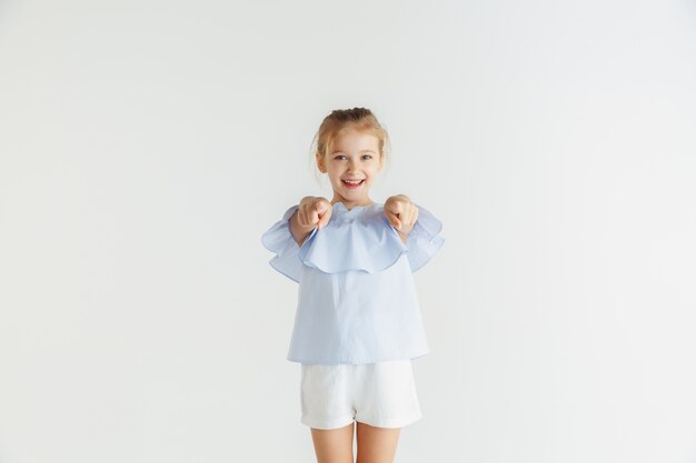 Niña sonriente con estilo posando en ropa casual aislado en la pared blanca. Modelo de mujer rubia caucásica. Emociones humanas, expresión facial, infancia. Señalar, elegir, sonreír.