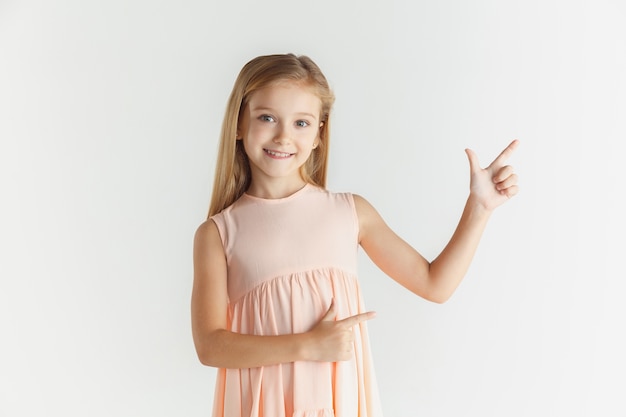 Niña sonriente con estilo posando en vestido aislado en la pared blanca. Modelo de mujer rubia caucásica. Emociones humanas, expresión facial, infancia. Apuntando a la barra espaciadora vacía.