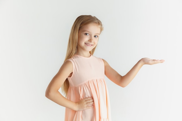 Niña sonriente con estilo posando en vestido aislado en la pared blanca. Modelo de mujer rubia caucásica. Emociones humanas, expresión facial, infancia. Mostrando en la barra espaciadora vacía.