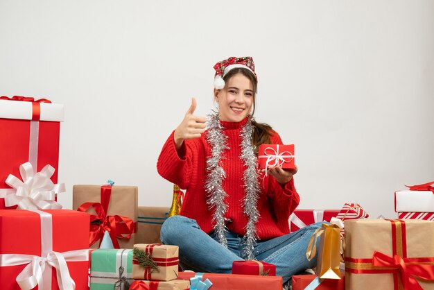 Niña sonriente con gorro de Papá Noel sosteniendo el presente y haciendo el pulgar hacia arriba firmar sentados alrededor de regalos en blanco
