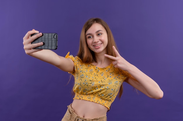 Niña sonriente haciendo el signo de la paz y tomando selfie en pared púrpura aislada