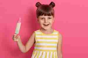 Foto gratuita niña sonriente con helado posando aislada sobre la pared de rosas con expresión alegre, sosteniendo hielo de agua en las manos, divertida niña con nudos, vestido de verano.