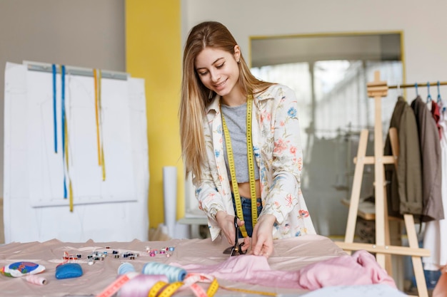 Niña sonriente joven soñadoramente usando tijeras para trabajar con tela en un taller de costura moderno