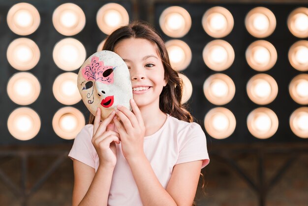 Niña sonriente con máscara veneciana en sus manos de pie delante de la luz de la etapa