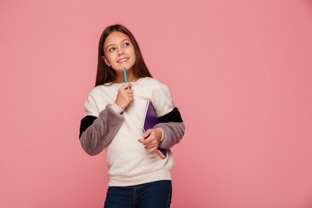 Niña sonriente mirando hacia arriba y pensando mientras sostiene un lápiz y libros