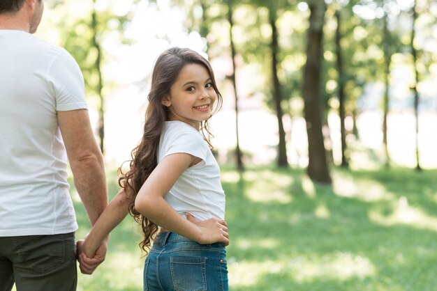 Niña sonriente mirando a cámara mientras camina en el parque con su padre