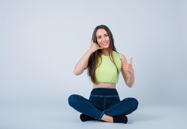 Foto gratuita niña sonriente muestra gesto de teléfono con la mano sentada en el suelo sobre fondo blanco.