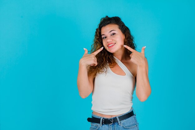 Niña sonriente muestra su sonrisa con los dedos índices sobre fondo azul.