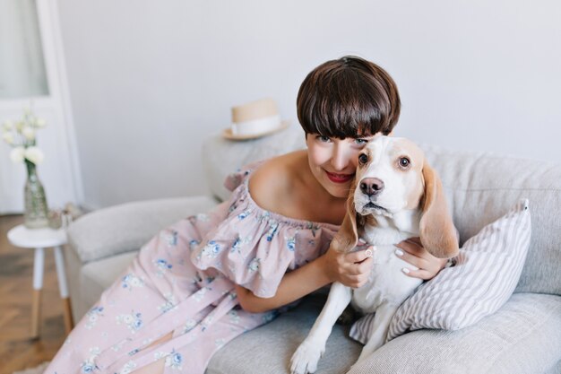 Niña sonriente de pelo castaño en vestido rosa jugando con su lindo perro beagle en casa