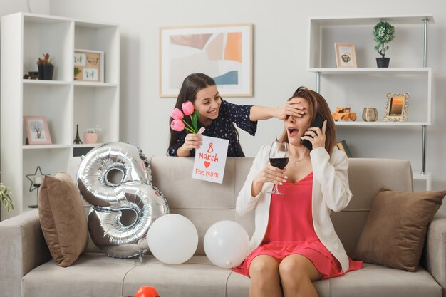 Niña sonriente de pie detrás del sofá sosteniendo la tarjeta de felicitación con flores cubrió los ojos de mamá con la mano en el día de la mujer feliz en la sala de estar