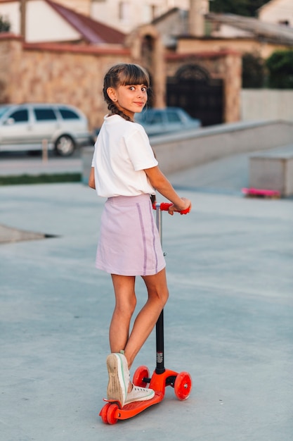 Foto gratuita niña sonriente de pie en empuje scooter mirando hacia atrás