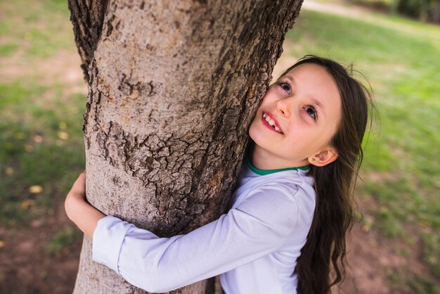 Niña sonriente que abraza el árbol en jardín