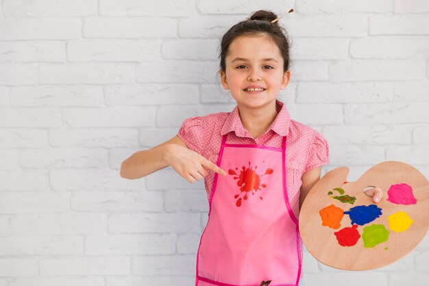 Niña sonriente que se coloca delante de la pared de ladrillo blanca que señala en la paleta colorida a disposición