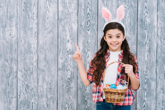 Niña sonriente que sostiene los huevos de Pascua en la cesta que señala el dedo que mira a la cámara