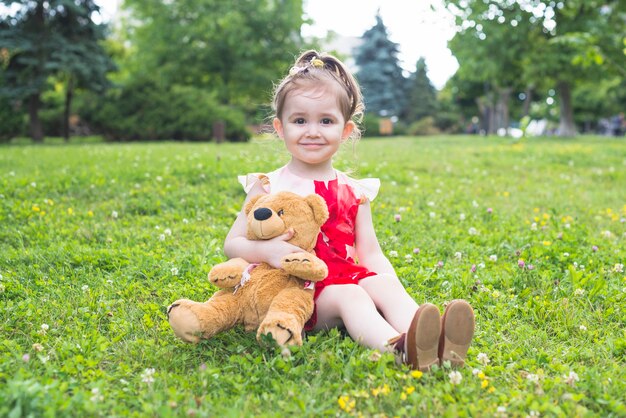 Niña sonriente que sostiene el oso de peluche que se sienta en la hierba verde