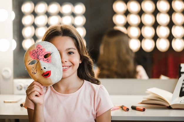 Foto gratuita niña sonriente sentada en el backstage sosteniendo la máscara delante de su cara