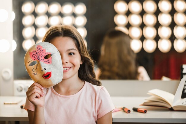 Niña sonriente sentada en el backstage sosteniendo la máscara delante de su cara