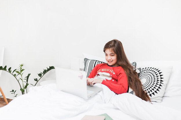 Niña sonriente sentada en la cama usando la computadora portátil en el dormitorio