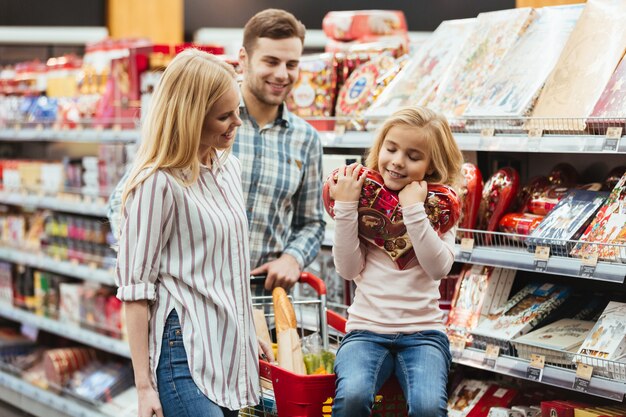 Niña sonriente sentada en un carrito de compras y elegir dulces con sus padres en el supermercado