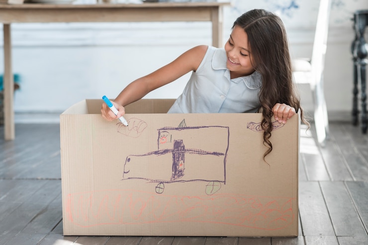 Foto gratuito niña sonriente sentada dentro de la caja de cartón dibujando con marcador