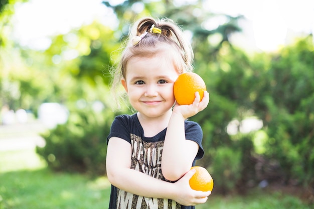 Niña sonriente sosteniendo dos naranjas frescas en el parque