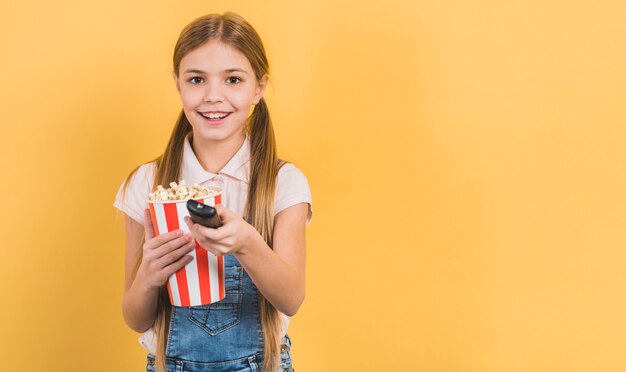 Niña sonriente sosteniendo palomitas de maíz en la mano cambiando el canal con control remoto sobre fondo amarillo