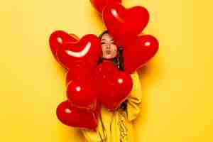 Foto gratuita niña sonriente en suéter amarillo dando un beso, mirando fuera de manojo de globos rojos.