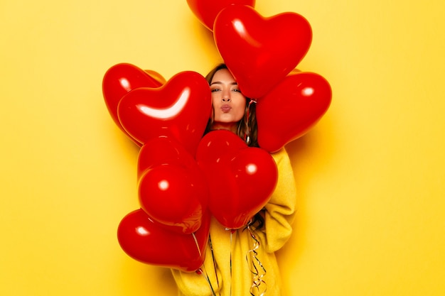 Niña sonriente en suéter amarillo dando un beso, mirando fuera de manojo de globos rojos.