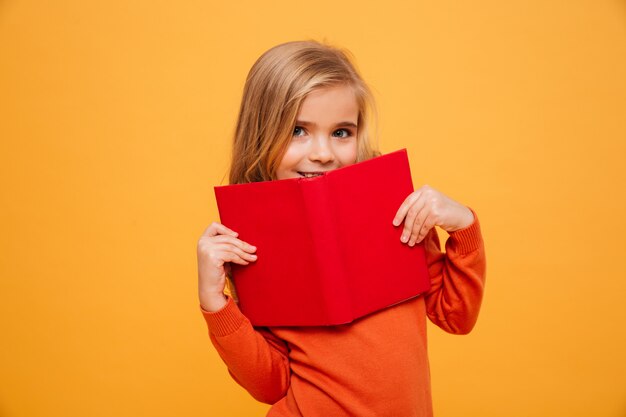 Niña sonriente en suéter escondiéndose detrás del libro y mirando a la cámara sobre naranja