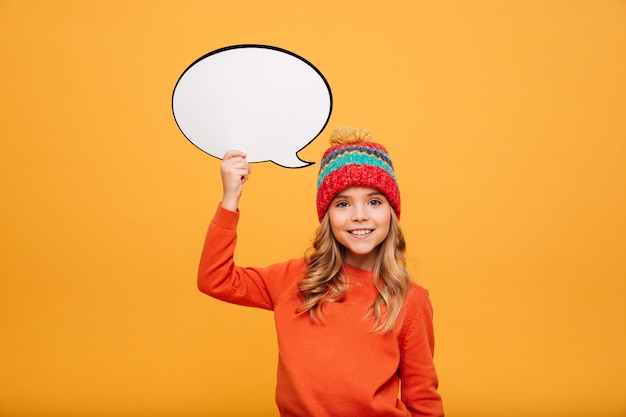 Niña sonriente en suéter y sombrero con discurso en blanco buble y mirando a la cámara sobre naranja