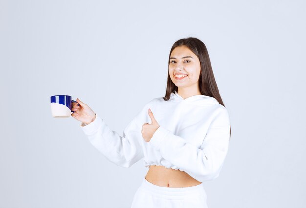 niña sonriente con una taza y mostrando un pulgar hacia arriba sobre fondo blanco-gris.