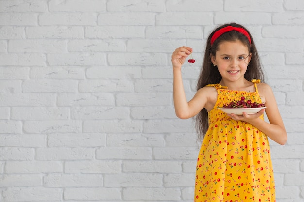 Niña sonriente, tenencia, cerezas, en, placa, posición, contra, pared de ladrillo