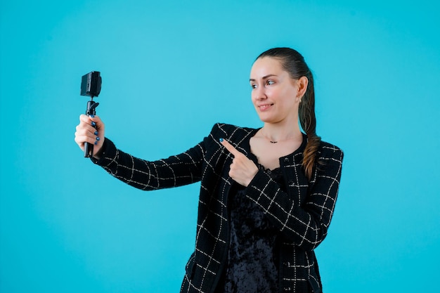 Niña sonriente se está tomando selfie con su mini cámara extendiendo el dedo índice a la cámara sobre fondo azul.