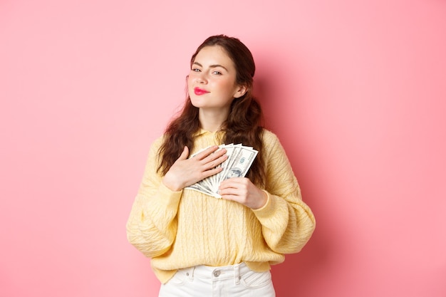 La niña sonriente se ve satisfecha y agradecida, abrazando billetes de un dólar, sosteniendo dinero y haciendo una cara engreída complacida, de pie contra el fondo rosa.