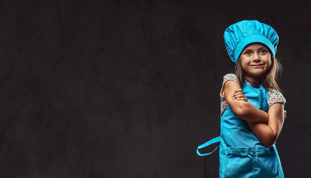 Niña sonriente vestida con uniforme de cocinero azul de pie con los brazos cruzados. Aislado en un fondo de textura oscura.