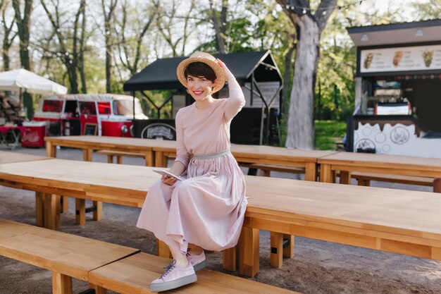 Niña sonriente en vestido largo y zapatos de moda sentado en la mesa de madera con teléfono móvil en la mano