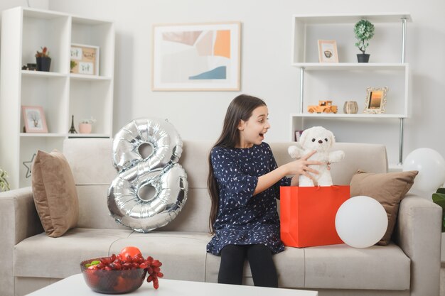 Niña sorprendida en el día de la mujer feliz sosteniendo y mirando a un oso de peluche sentado en el sofá en la sala de estar