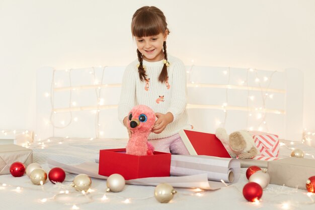 Niña sorprendida emocionada con suéter blanco sosteniendo un peluche rosa en las manos, mirando su regalo con ojos grandes, sentada en la cama con decoración navideña y guirnalda.