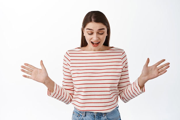 La niña sorprendida encuentra algo debajo, mirando hacia abajo y regocijándose con la cara de asombro feliz, extiende las manos hacia los lados como si estuviera viendo algo inesperado, de pie sobre el fondo blanco