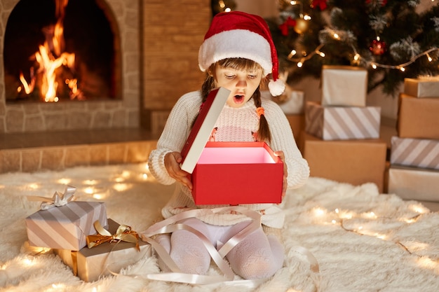 Niña sorprendida extremadamente emocionada con suéter blanco y sombrero de santa claus, abre la caja de regalo con algo brillante, sentada en el piso cerca del árbol de Navidad, cajas de regalo y chimenea.