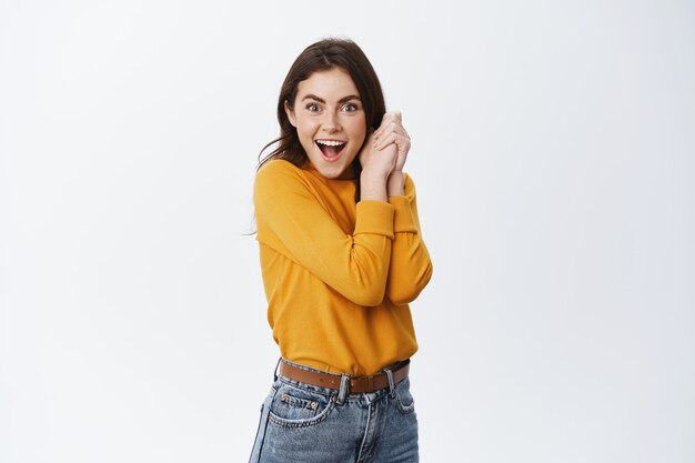 Niña sorprendida recibe una oferta interesante, jadeando sorprendida y feliz, sosteniendo las manos apretadas contra el cuerpo y sonriendo al frente, mirando al premio, pared blanca