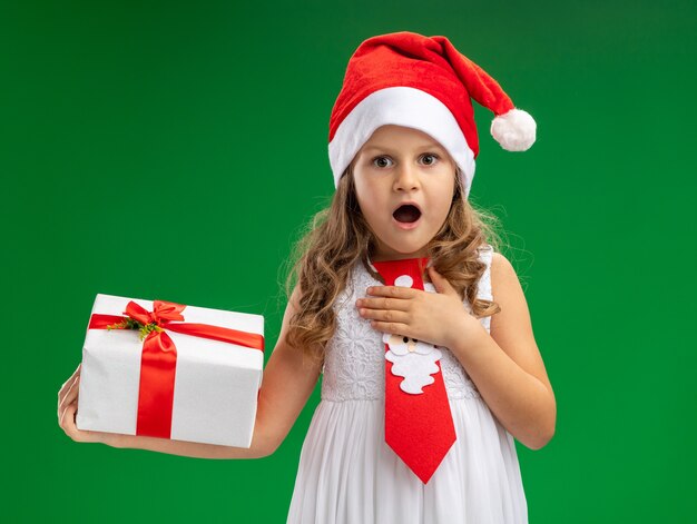 Niña sorprendida con sombrero de navidad con corbata sosteniendo caja de regalo poniendo la mano sobre sí misma aislada sobre fondo verde