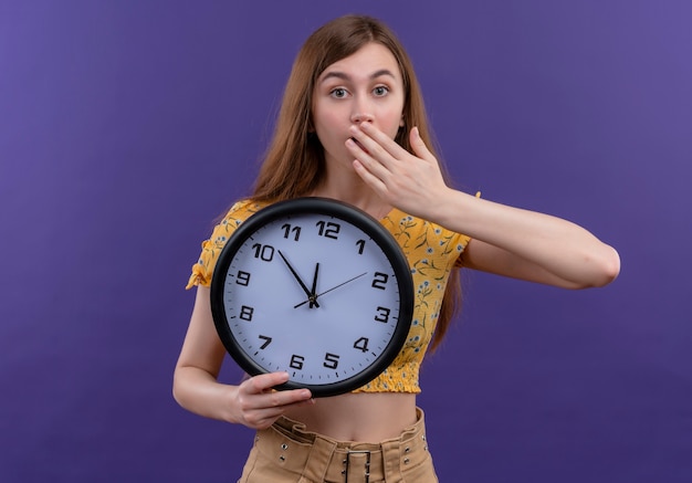 Niña sorprendida sosteniendo el reloj y poniendo la mano en la boca en la pared púrpura aislada