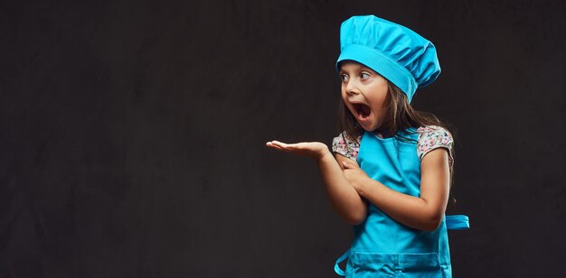 Niña sorprendida vestida de cocinero azul posando en un estudio. Aislado en un fondo de textura oscura.