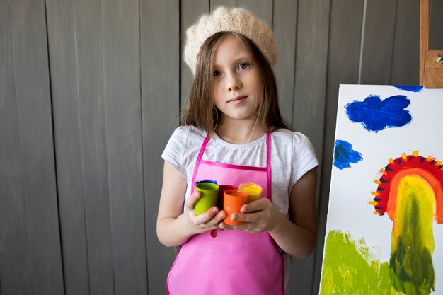 Foto gratuita una niña sosteniendo botellas de pintura de colores en la mano de pie cerca de la base