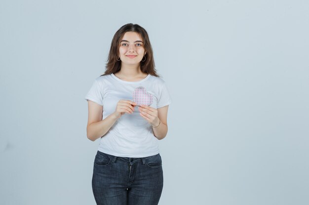 Niña sosteniendo una caja de regalo mientras mira la caja en camiseta, jeans y luce alegre, vista frontal.